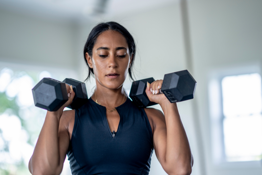 A close up of a woman holding dumbbells in each hand and resting them on her shoulders