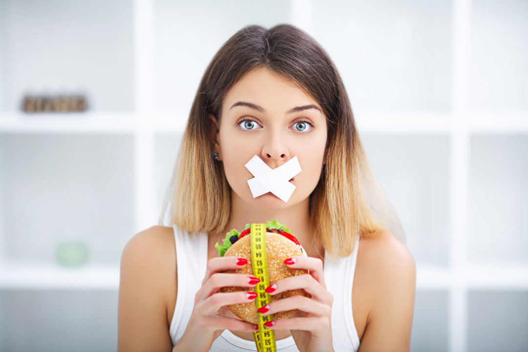 A woman with tape over her mouth, holding a burger with a measuring tape around it