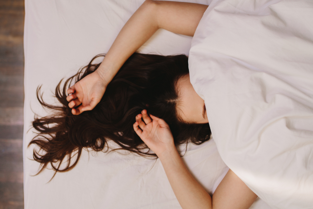 A woman with long dark hair asleep in bed with the covers over her face